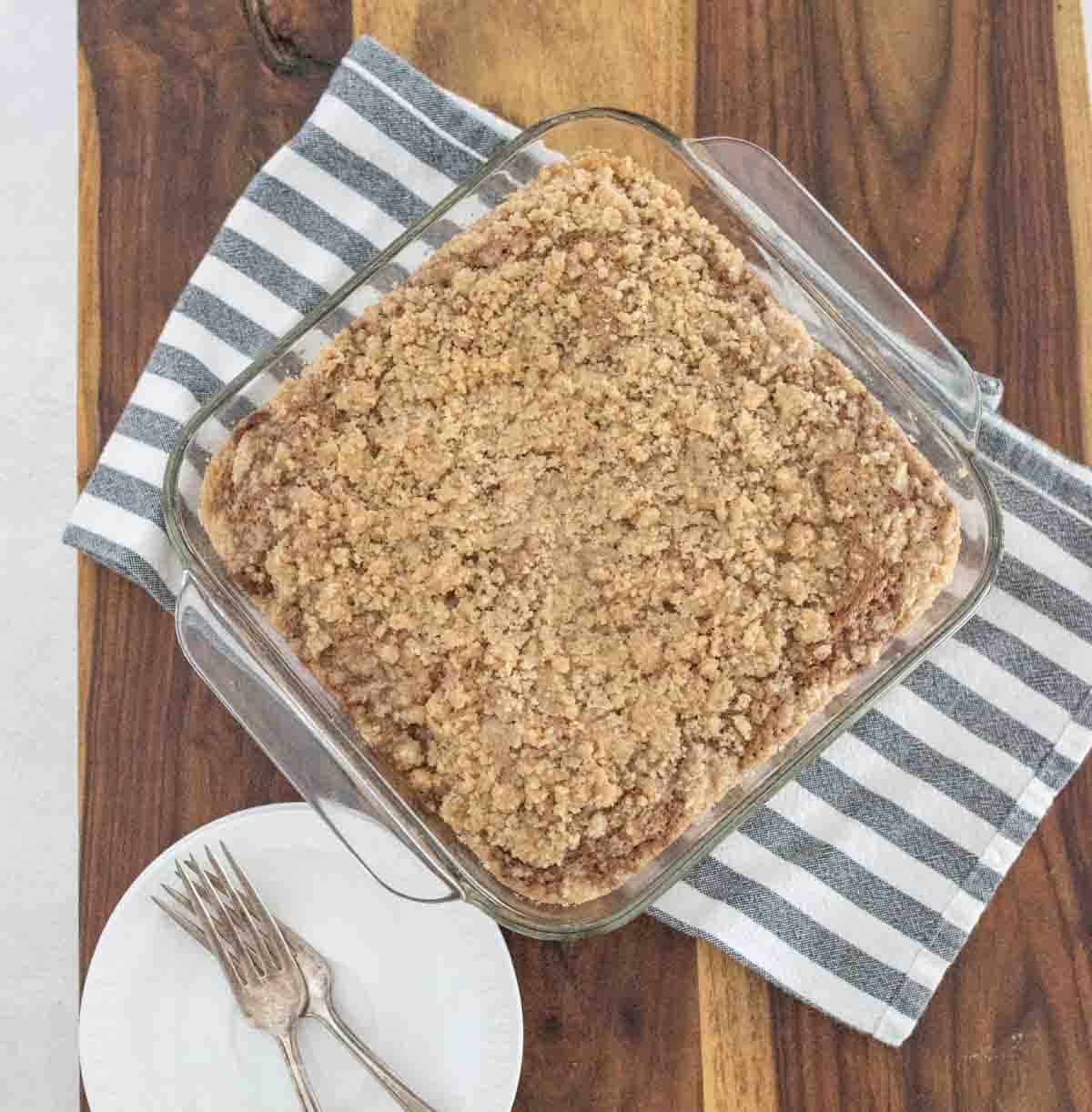 A square glass baking dish filled with crumb-topped applesauce coffee cake sits on a striped towel on a wooden table. Nearby, a white plate holds two forks.