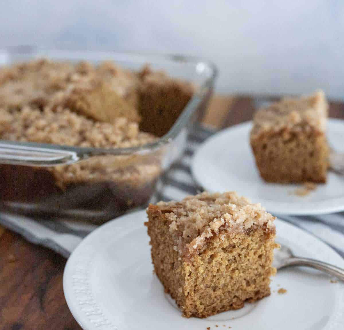 A close-up of two slices of crumb-topped applesauce coffee cake on white plates, with a glass baking dish of more coffee cake in the background. A fork rests beside one of the slices.
