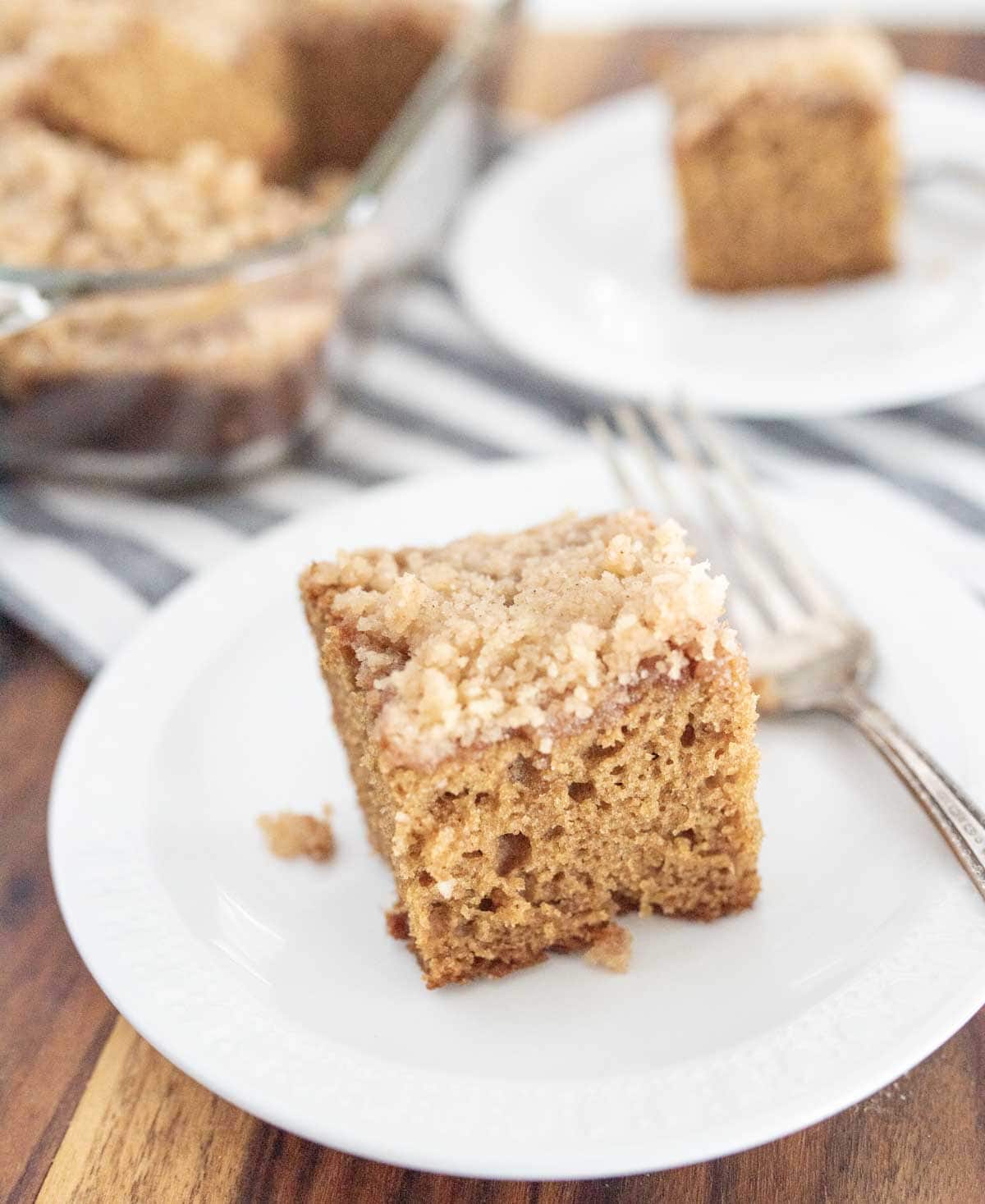A slice of crumb-topped applesauce coffee cake sits on a white plate with a fork, with another plate and the rest of the cake visible in the background on a striped cloth.