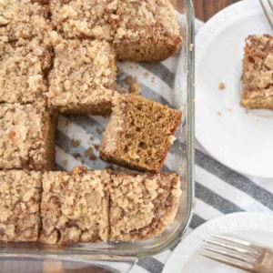 A glass baking dish filled with sliced pieces of crumb-topped applesauce coffee cake, with two pieces served on white plates beside it, each with a fork. The dish sits on a striped cloth on a wooden surface.