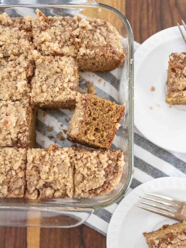 A glass baking dish filled with sliced pieces of crumb-topped applesauce coffee cake, with two pieces served on white plates beside it, each with a fork. The dish sits on a striped cloth on a wooden surface.