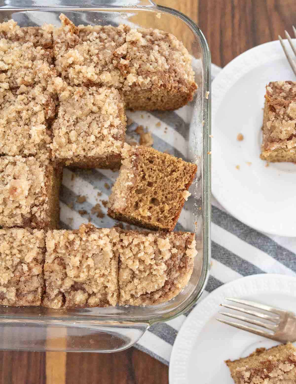 Overhead view of a glass baking dish with sliced crumb-topped coffee cake. Two small square pieces are served on white plates with forks, placed on a striped cloth on a wooden table.