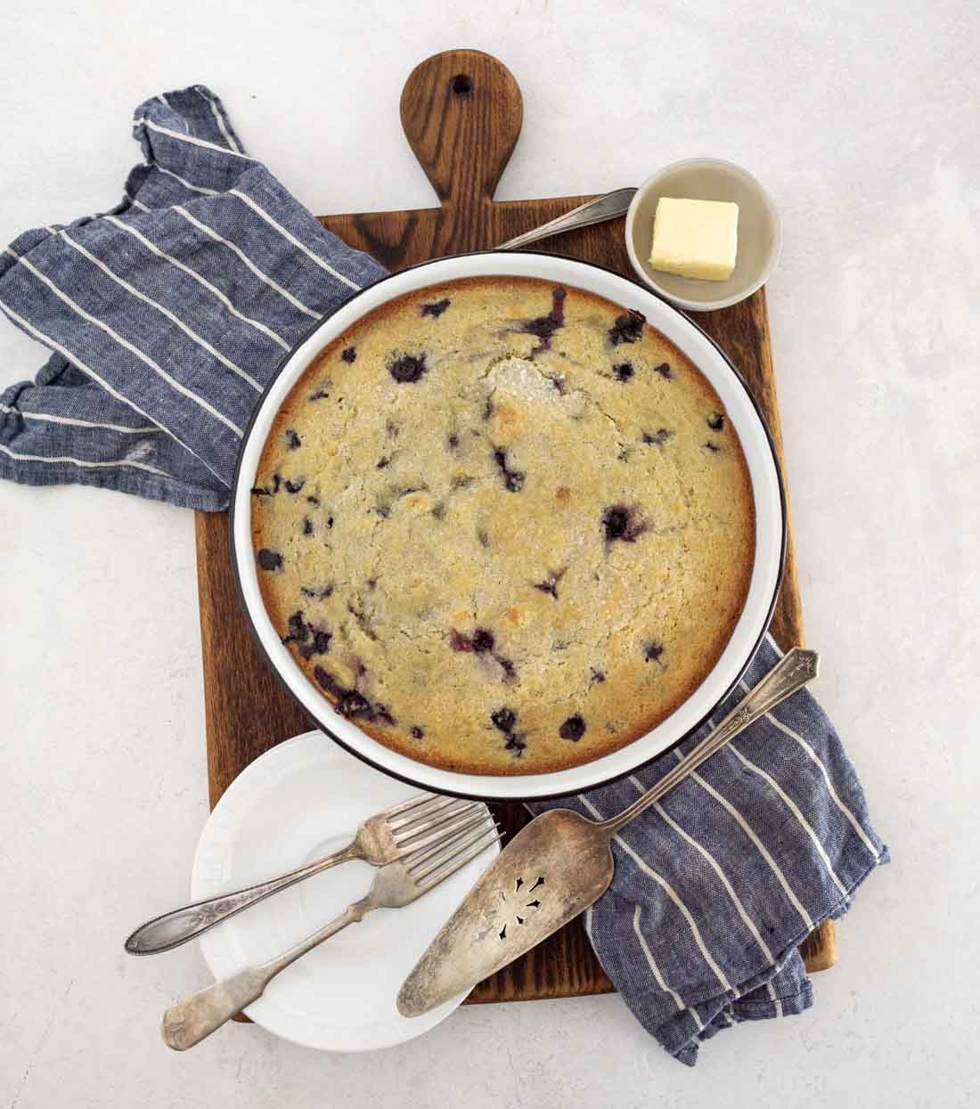 A round blueberry cornbread in a baking dish sits on a wooden board, surrounded by a small plate, fork, pie server, butter dish, knife, and two striped blue napkins. This blueberry cornbread makes for a cozy, inviting table setting.