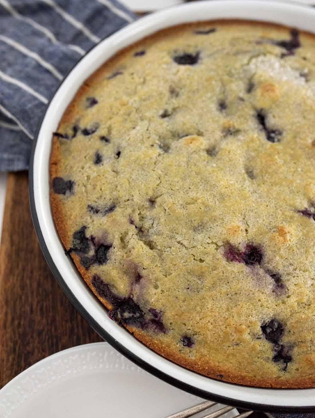 A freshly baked blueberry cornbread cake in a round white pan, with a golden-brown top and visible blueberries. A striped cloth is partially visible beside the pan on a wooden surface.