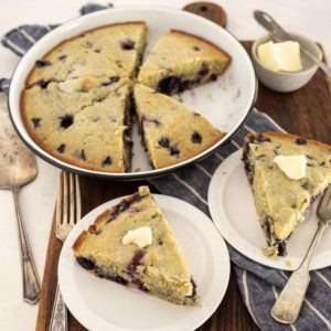 A round blueberry cornbread, sliced and served on white plates; each slice of the blueberry cornbread is topped with a pat of butter. The plates rest on a wooden board alongside a fork, napkin, and extra butter.