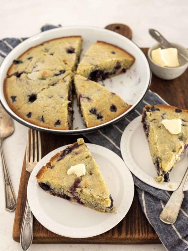 A round blueberry cornbread, sliced and served on white plates; each slice of the blueberry cornbread is topped with a pat of butter. The plates rest on a wooden board alongside a fork, napkin, and extra butter.
