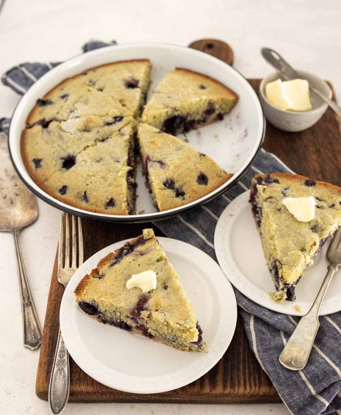 A round blueberry cornbread, sliced and served on white plates; each slice of the blueberry cornbread is topped with a pat of butter. The plates rest on a wooden board alongside a fork, napkin, and extra butter.