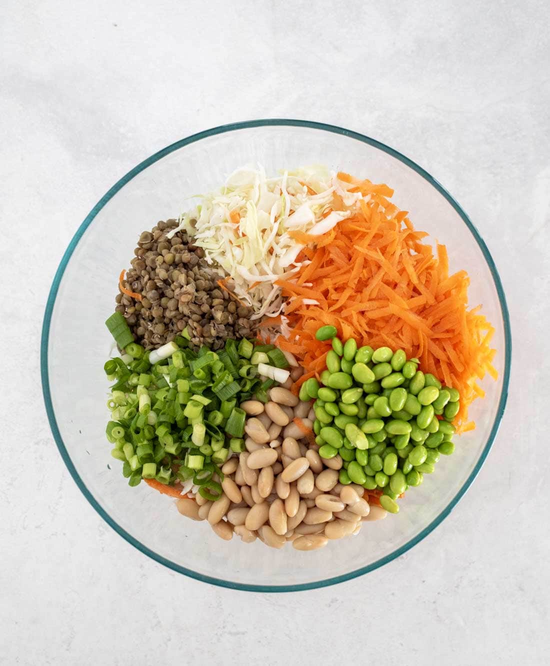A glass bowl containing shredded cabbage, grated carrots, green onions, edamame, white beans, and lentils, arranged in separate sections on a white surface.