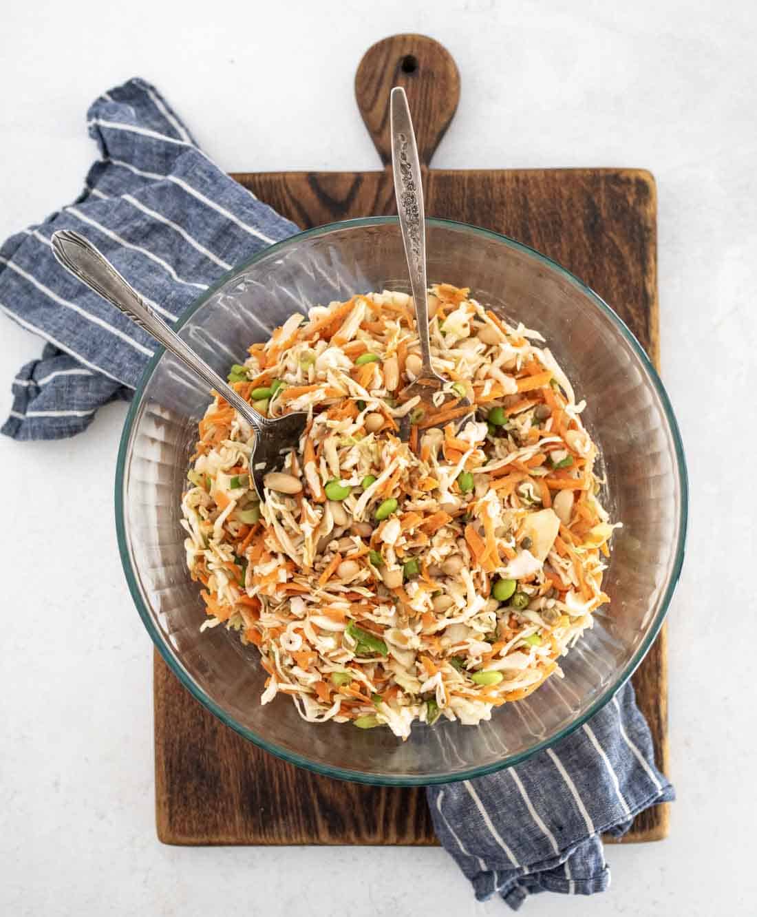 A glass bowl filled with a colorful salad of shredded cabbage, carrots, edamame, and nuts, with two serving utensils. The bowl sits on a wooden board with a blue striped cloth underneath.