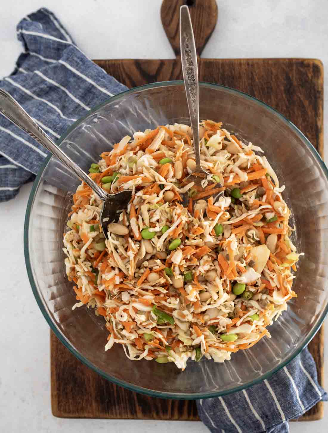 A glass bowl filled with shredded cabbage, carrots, edamame, and nuts, with two serving utensils. The bowl sits on a wooden board next to a blue and white striped cloth.