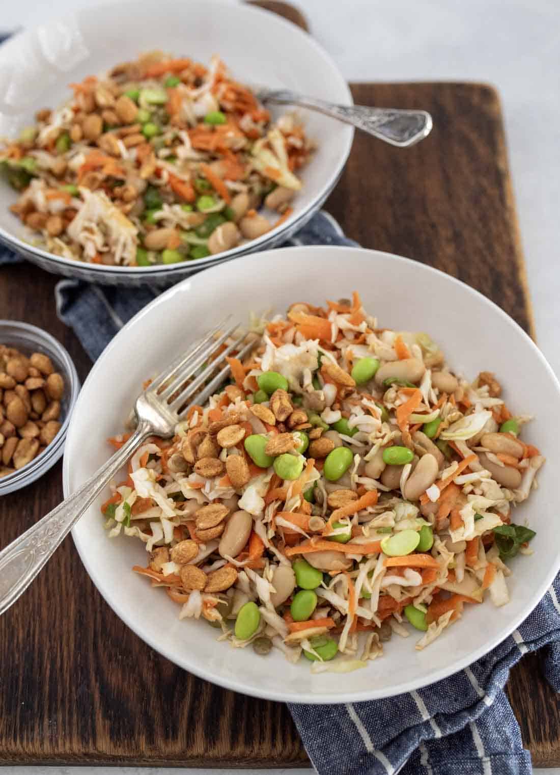 Two white bowls filled with a colorful salad of shredded cabbage, carrots, edamame, peanuts, and beans sit on a wooden board. A small bowl of peanuts and two forks are also visible.