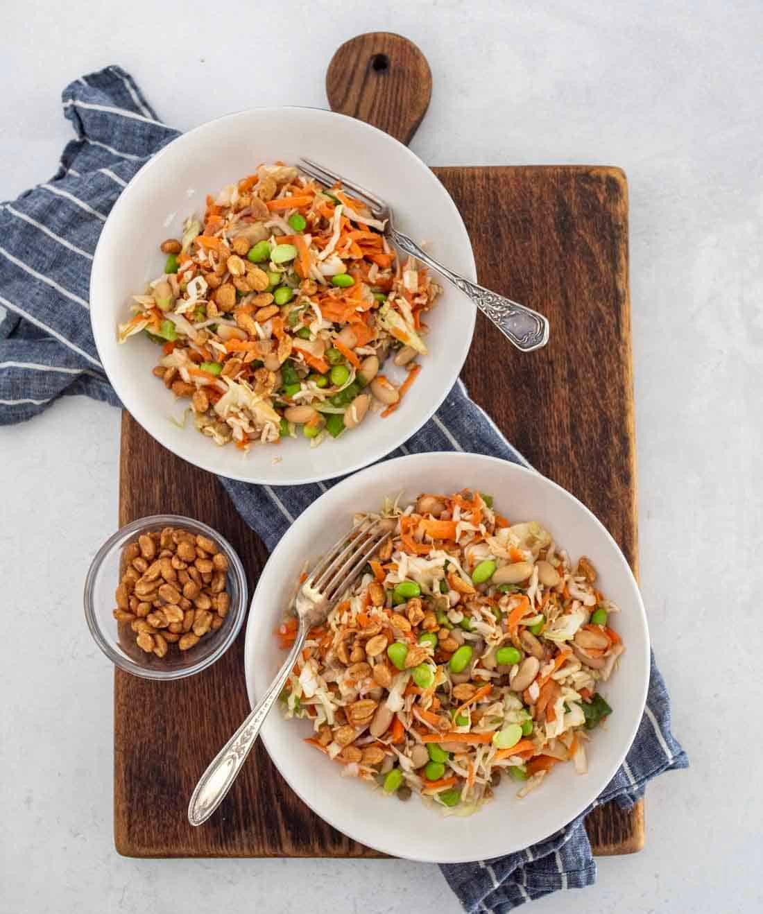 Two white bowls of colorful salad with shredded carrots, cabbage, edamame, and nuts are placed on a wooden board with a blue cloth. A small bowl of nuts sits beside the salads, each with a fork.