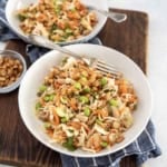 Two white bowls filled with colorful salad containing shredded cabbage, carrots, edamame, white beans, and chopped peanuts, placed on a wooden board with a small bowl of peanuts and a blue checked cloth.