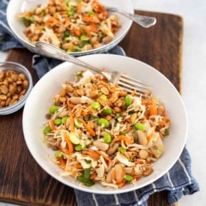 Two white bowls filled with colorful salad containing shredded cabbage, carrots, edamame, white beans, and chopped peanuts, placed on a wooden board with a small bowl of peanuts and a blue checked cloth.
