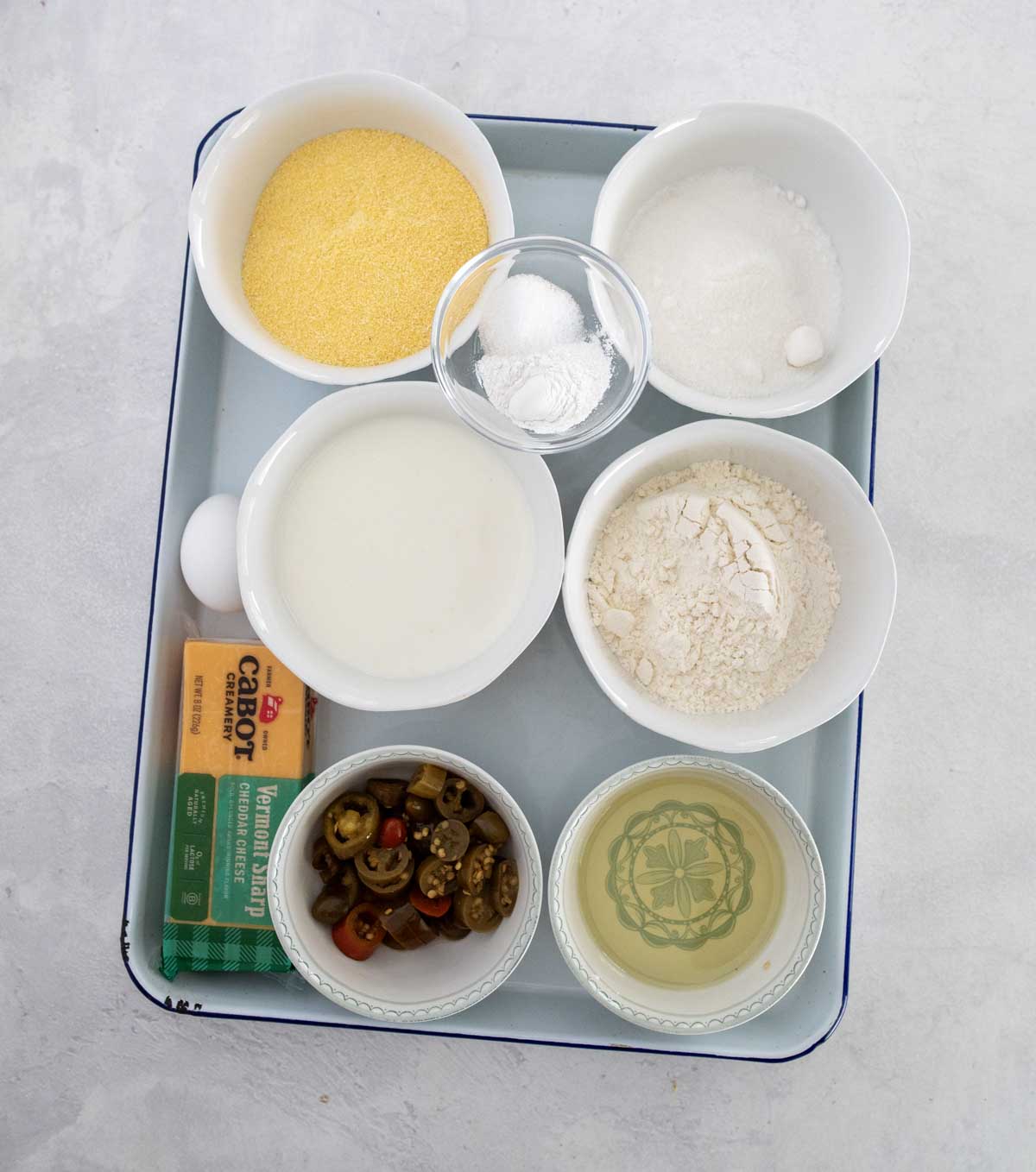 A tray with bowls containing cornmeal, sugar, flour, baking powder, milk, an egg, sliced jalape&ntilde;os, a stick of butter, and a small bowl of oil, arranged on a light surface.