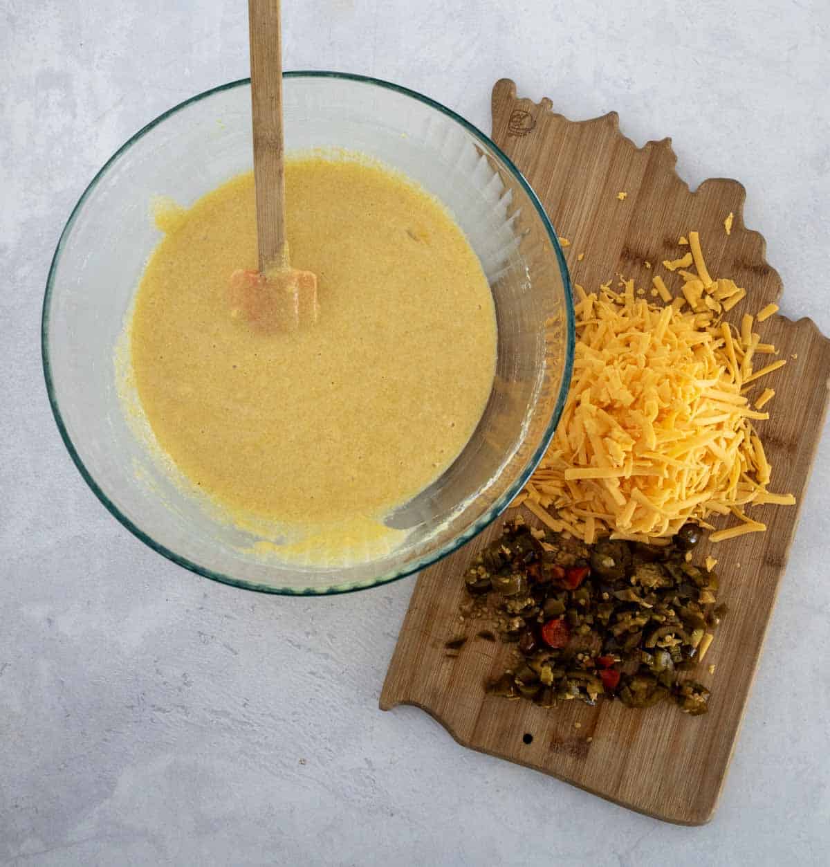 A glass bowl with cornmeal batter and a spatula next to a wooden board holding shredded cheddar cheese and chopped jalape&ntilde;os on a light surface.