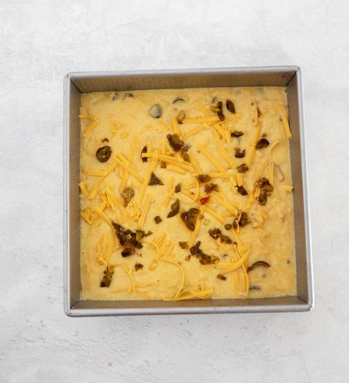 A square metal baking pan filled with unbaked cornbread batter, topped with shredded cheddar cheese and chopped jalape&ntilde;os, on a light-colored surface.