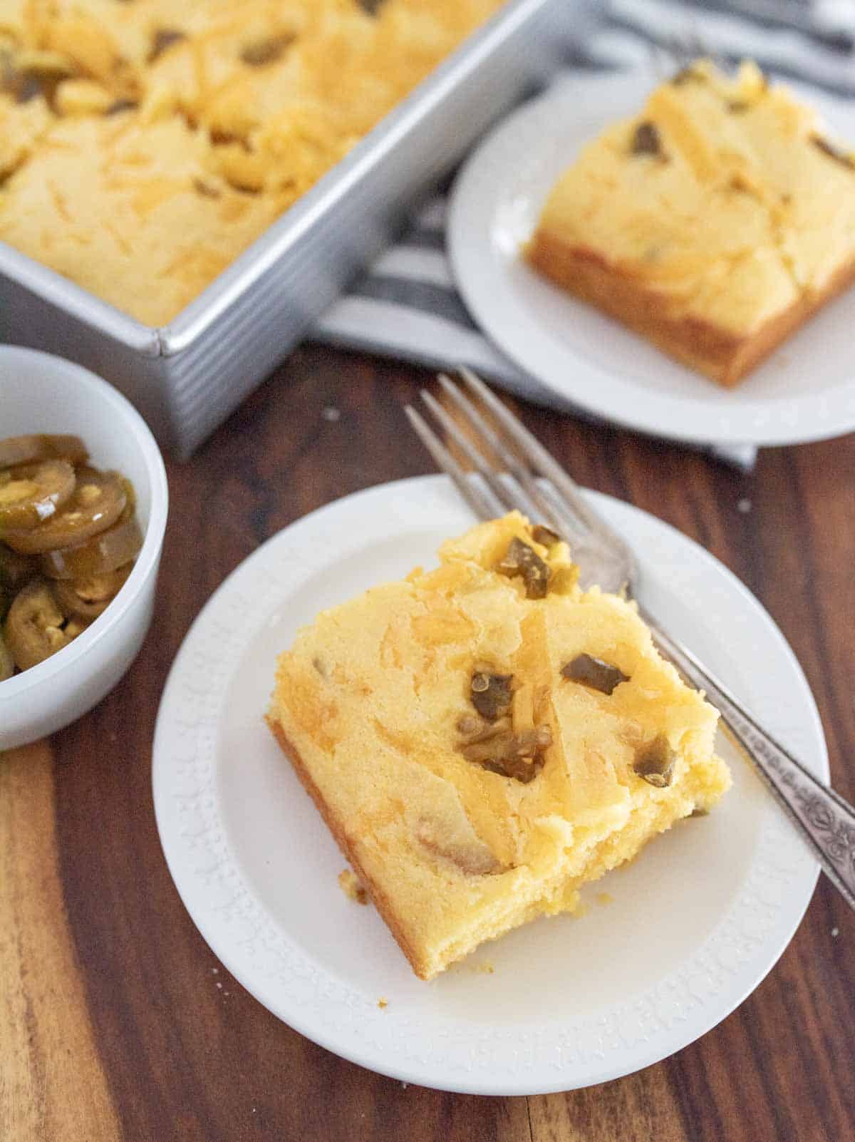 A slice of cornbread topped with jalape&ntilde;os sits on a white plate with a fork. A pan of more cornbread and another plate with a slice are in the background, along with a small bowl of jalape&ntilde;os.