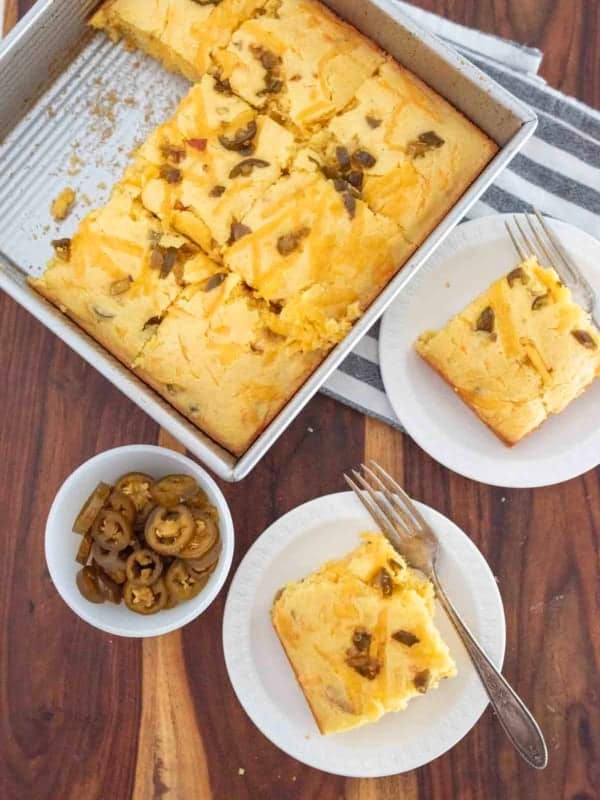 A square baking pan of cheddar and jalapeno cornbread sits on a wooden table. Two plates each hold a cheesy slice with a fork, and a small bowl contains pickled jalape&ntilde;o slices.
