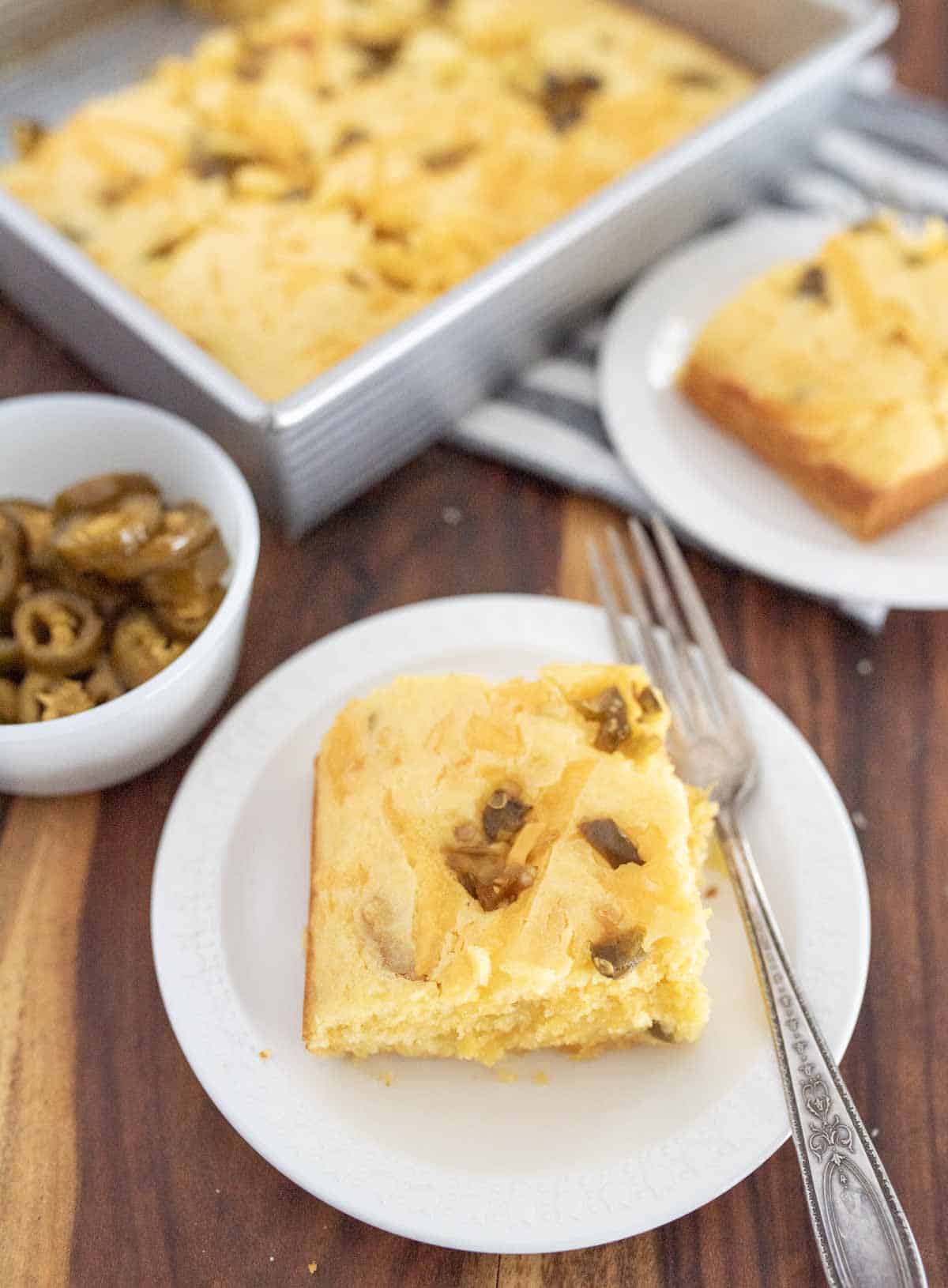 A slice of cornbread with jalape&ntilde;os on a white plate with a fork, next to a bowl of sliced jalape&ntilde;os and a pan of cornbread, all on a wooden table.