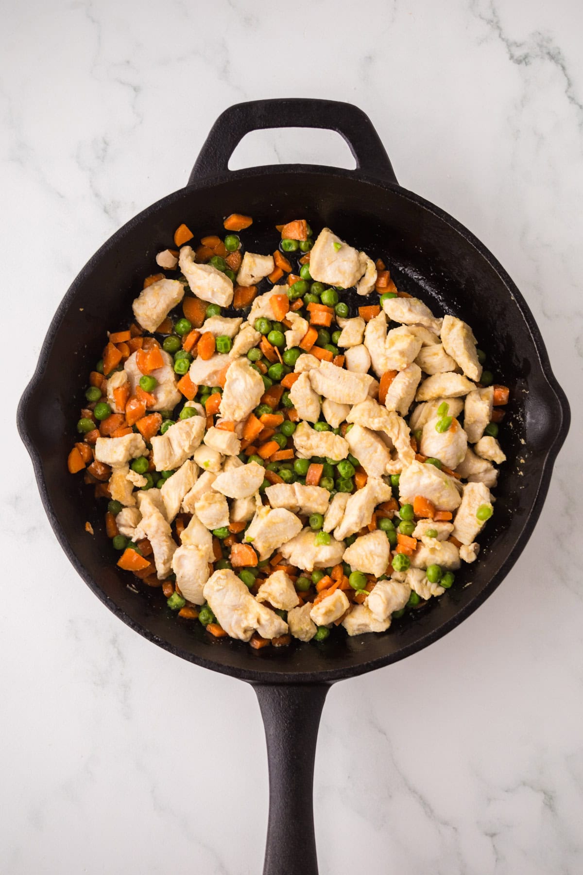 A cast iron skillet filled with cooked diced chicken, carrots, and peas on a white marble surface—perfect ingredients for making chicken fried rice.