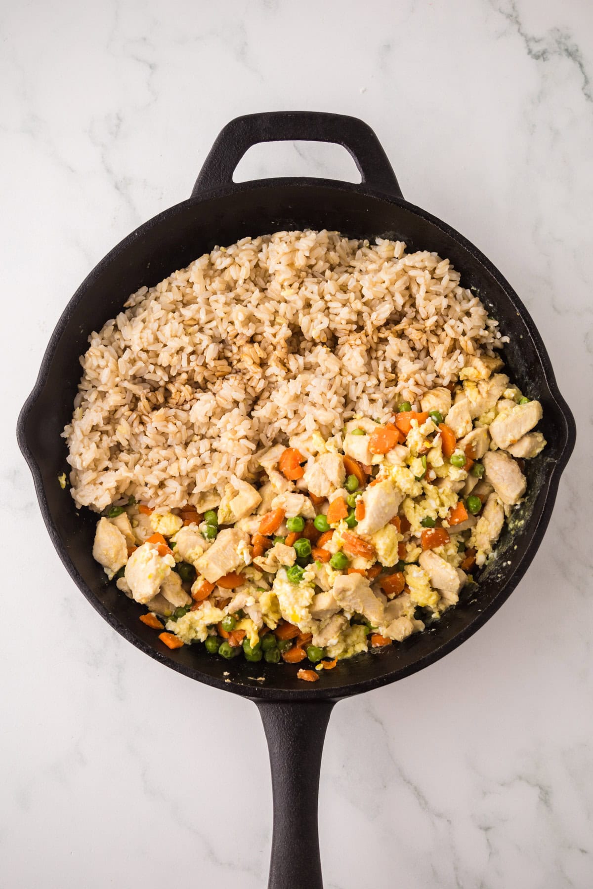 A black skillet on a marble surface, perfect for making chicken fried rice, filled with cooked brown rice on one side and a mix of diced chicken, peas, carrots, and scrambled eggs on the other.