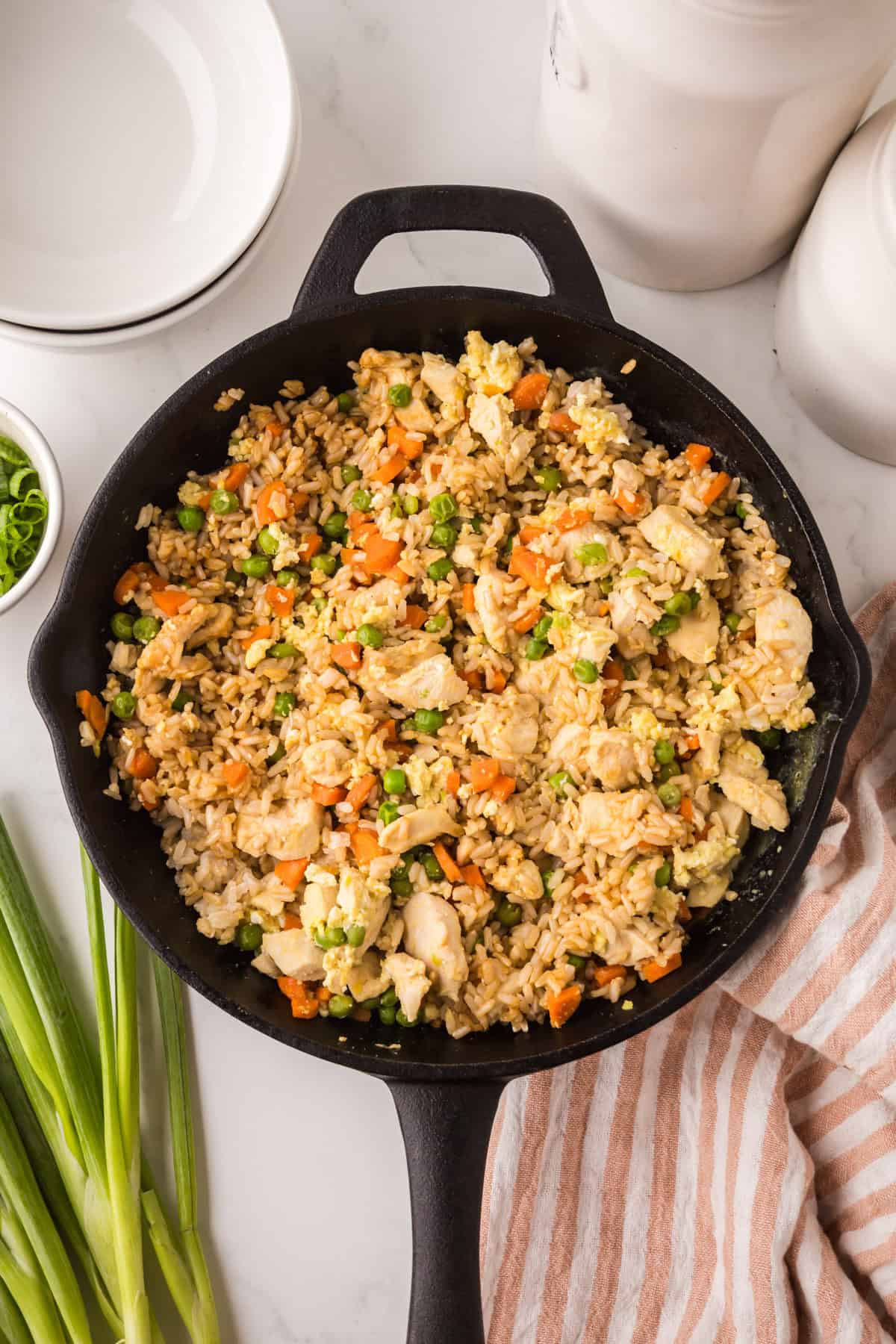 A skillet filled with chicken fried rice, made by making chicken fried rice with pieces of chicken, scrambled eggs, peas, carrots, and rice. Green onions, stacked plates, and a striped towel are nearby on a marble surface.