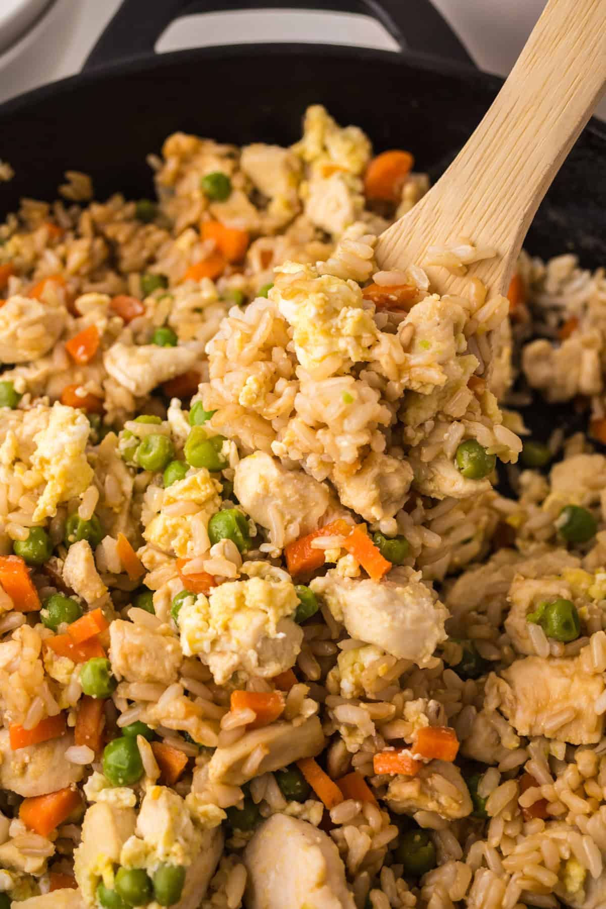 A close-up of making chicken fried rice in a skillet, with pieces of chicken, scrambled eggs, peas, and carrots. A wooden spoon is scooping up a portion.