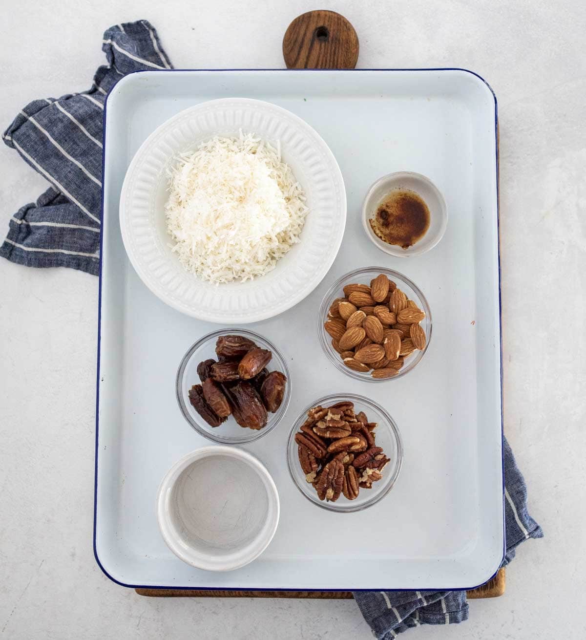 A white tray holds a bowl of shredded coconut, four small glass bowls with vanilla extract, whole almonds, chopped dates, and pecans, along with an empty small bowl. A striped kitchen towel lies beside the tray.