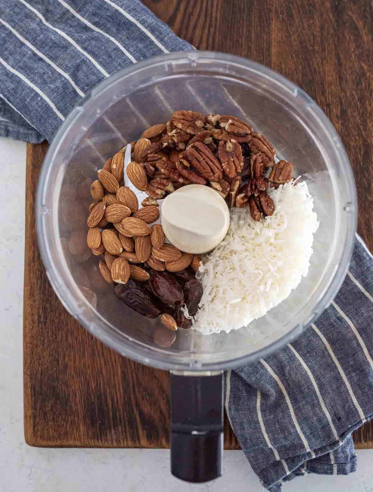 A food processor bowl contains whole almonds, pecans, pitted dates, and shredded coconut, set on a wooden cutting board next to a blue and white striped kitchen towel.