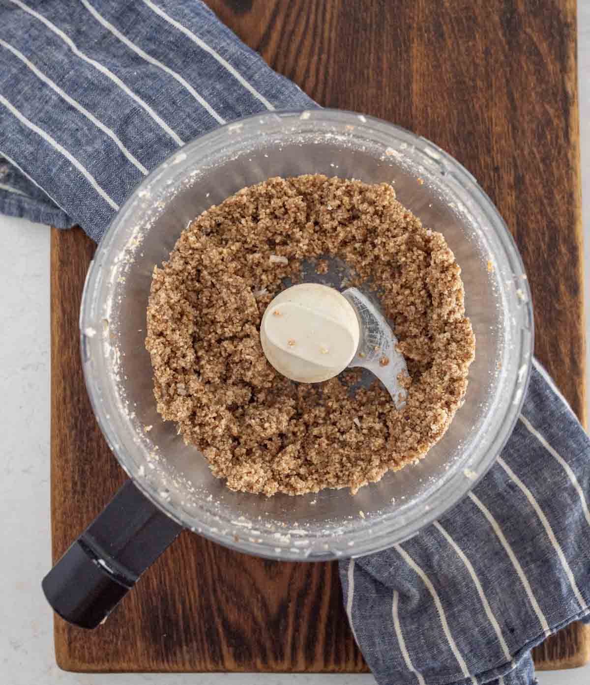 A food processor with a coarse, crumbly mixture inside sits on a wooden board with a blue and white striped kitchen towel underneath.