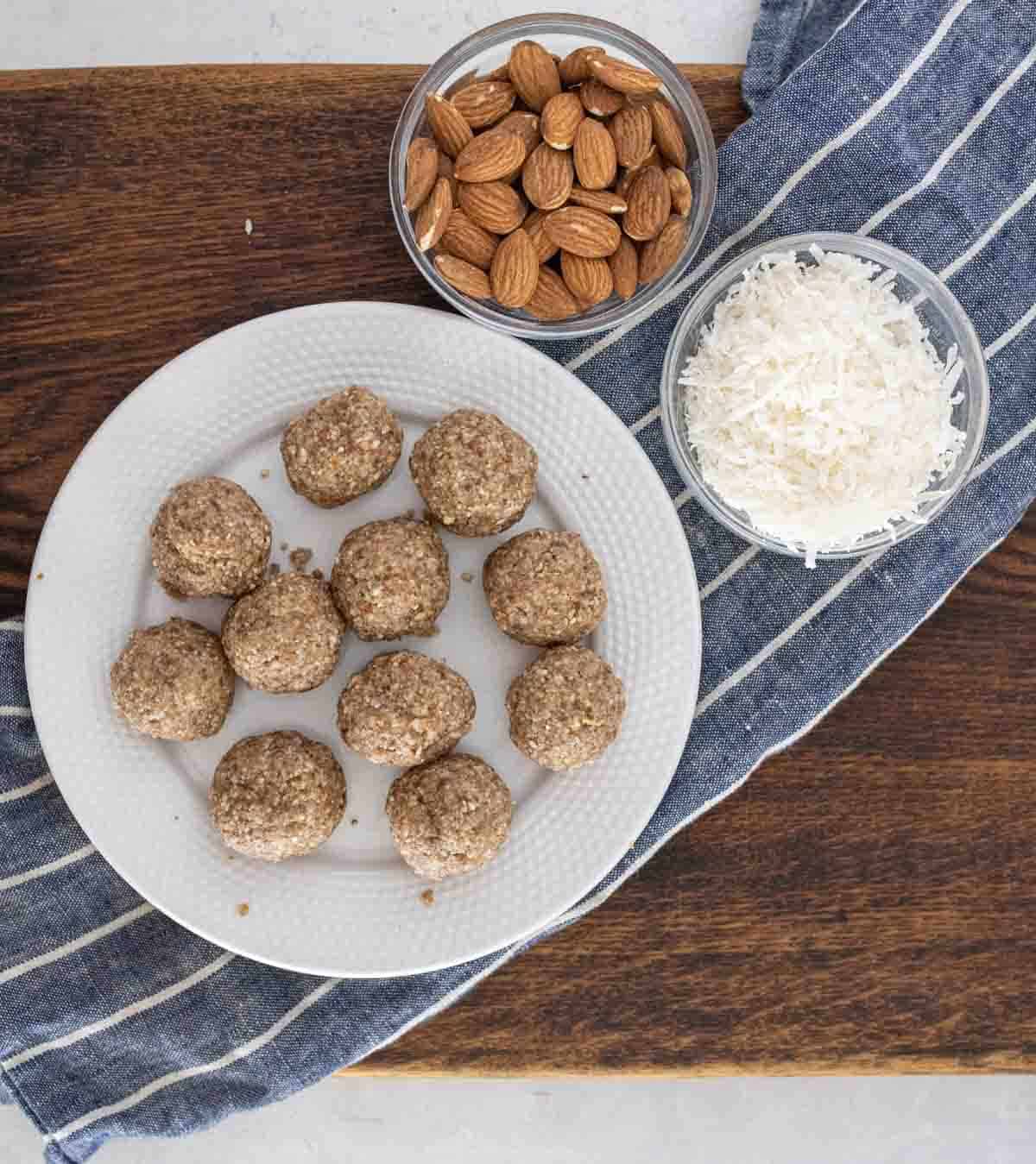 A white plate with ten almond energy balls sits on a wooden surface near two bowls&mdash;one with whole almonds and one with shredded coconut&mdash;on top of a blue-and-white striped cloth.