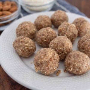 A white plate with several round, brown energy balls on it. In the background are small glass bowls containing shredded coconut and whole almonds, sitting on a wooden surface.