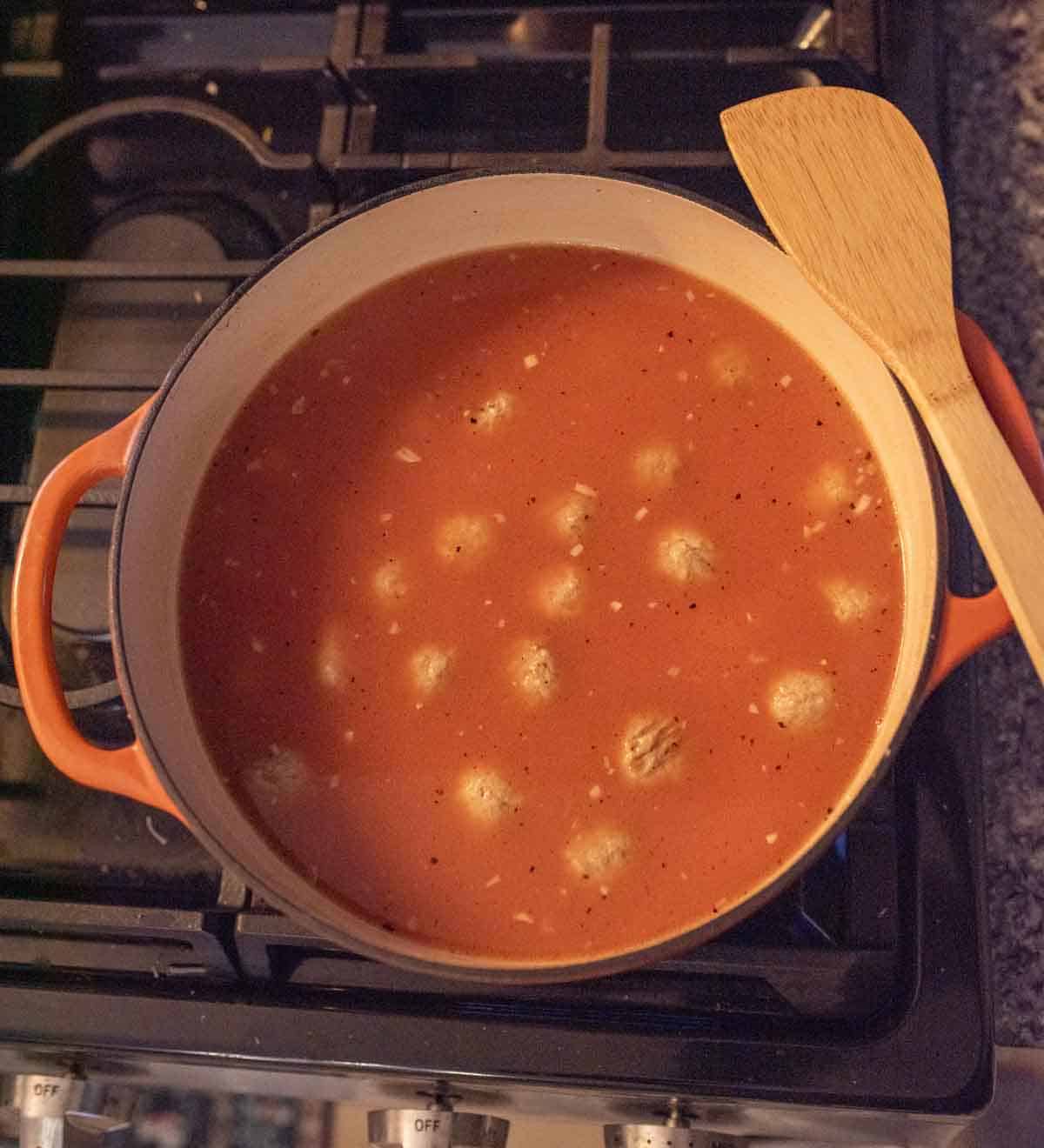 A pot of tomato soup with meatballs sits on a stove, with a wooden spatula resting on the edge. The soup is simmering, and small pieces of onion are visible in the red broth.