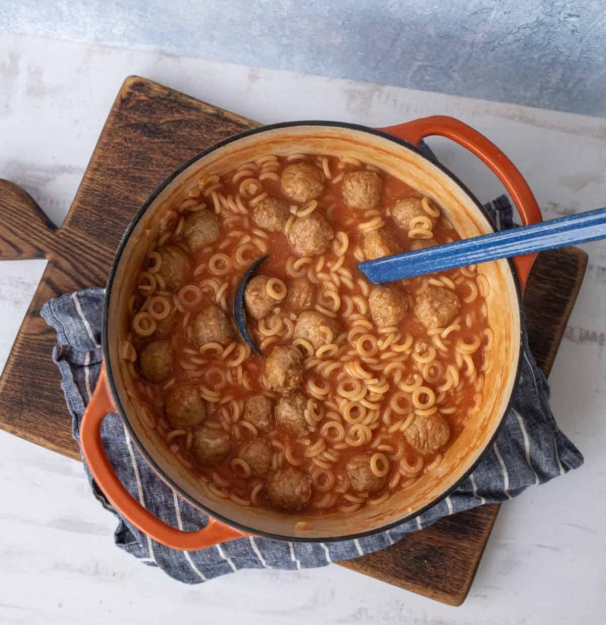 A red Dutch oven filled with tomato soup, meatballs, and ring-shaped pasta sits on a striped cloth atop a wooden cutting board. A blue-handled ladle rests inside the pot.