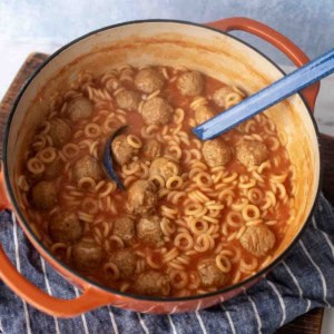 A red Dutch oven filled with ring-shaped pasta, meatballs, and tomato sauce, with a blue ladle resting inside. The pot sits on a striped cloth on a wooden surface.