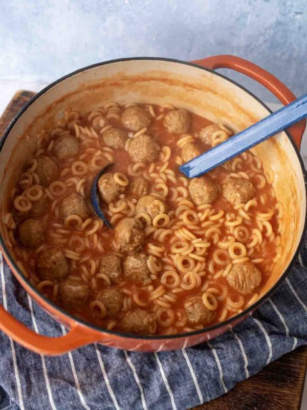 A red Dutch oven filled with ring-shaped pasta, meatballs, and tomato sauce, with a blue ladle resting inside. The pot sits on a striped cloth on a wooden surface.