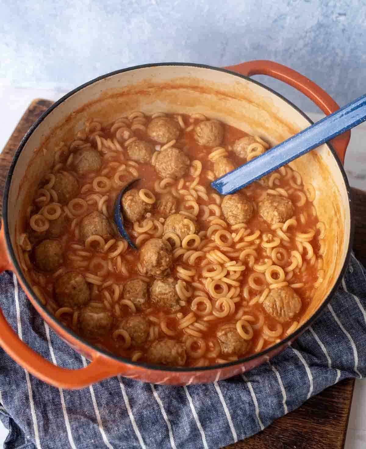 A red Dutch oven filled with ring-shaped pasta, meatballs, and tomato sauce, with a blue ladle resting inside. The pot sits on a striped cloth on a wooden surface.