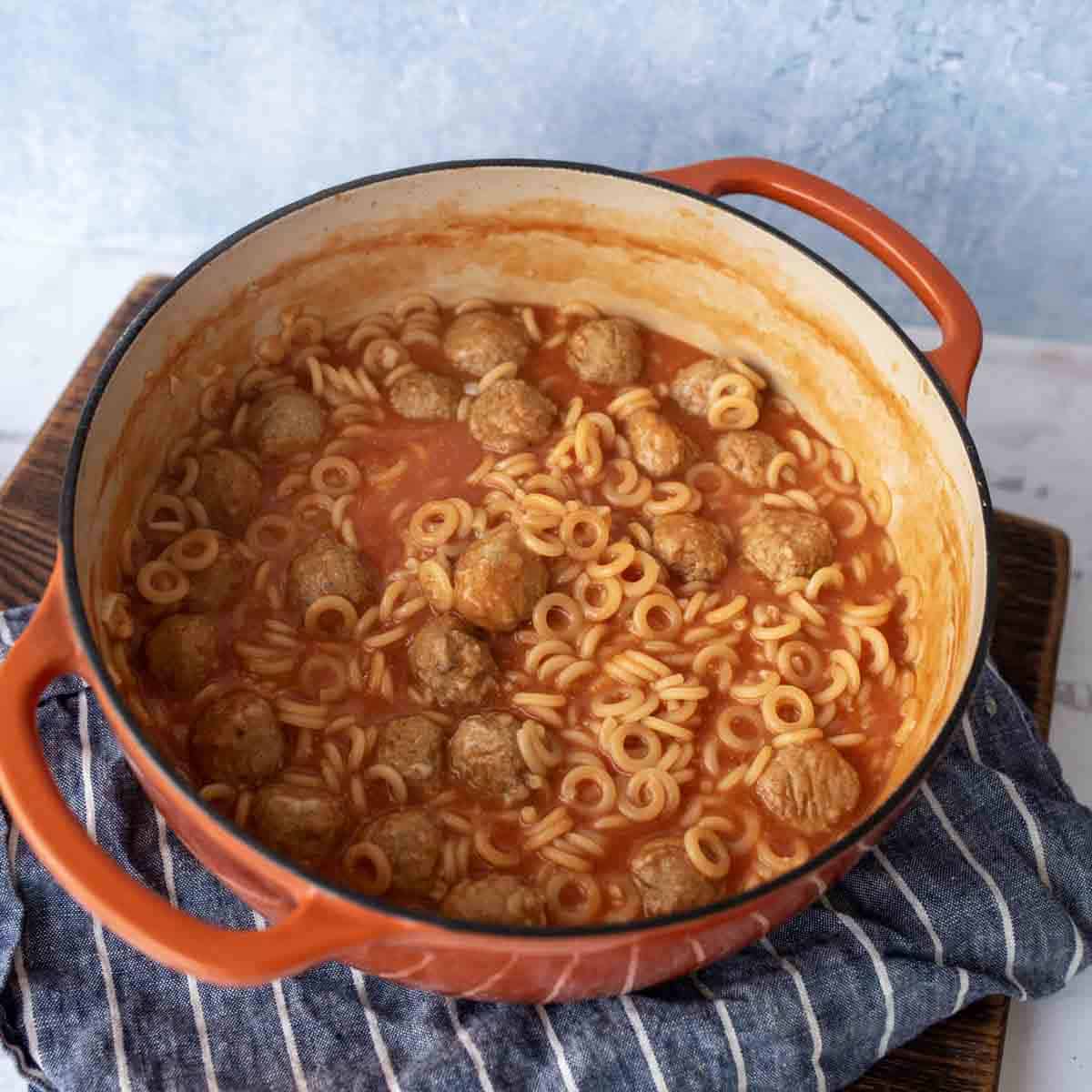 An orange pot filled with tomato soup, meatballs, and ring-shaped pasta sits on a blue and white striped cloth against a light blue background.