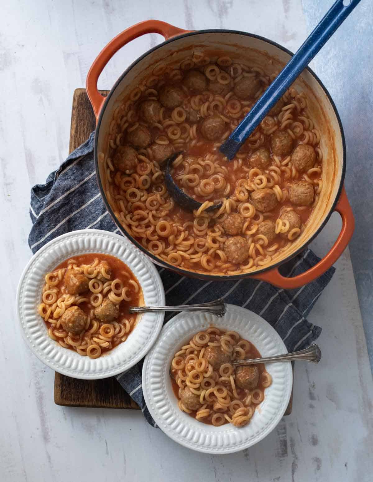 A pot of pasta with meatballs in tomato sauce sits on a blue-striped cloth, with two white bowls of the same dish and metal spoons beside it, ready to serve.