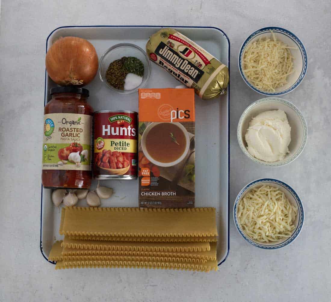 A tray with ingredients for lasagna soup in slow cooker: lasagna noodles, garlic, onion, marinara sauce, diced tomatoes, chicken broth, ground sausage, ricotta cheese, and two bowls of shredded cheese on a white surface.