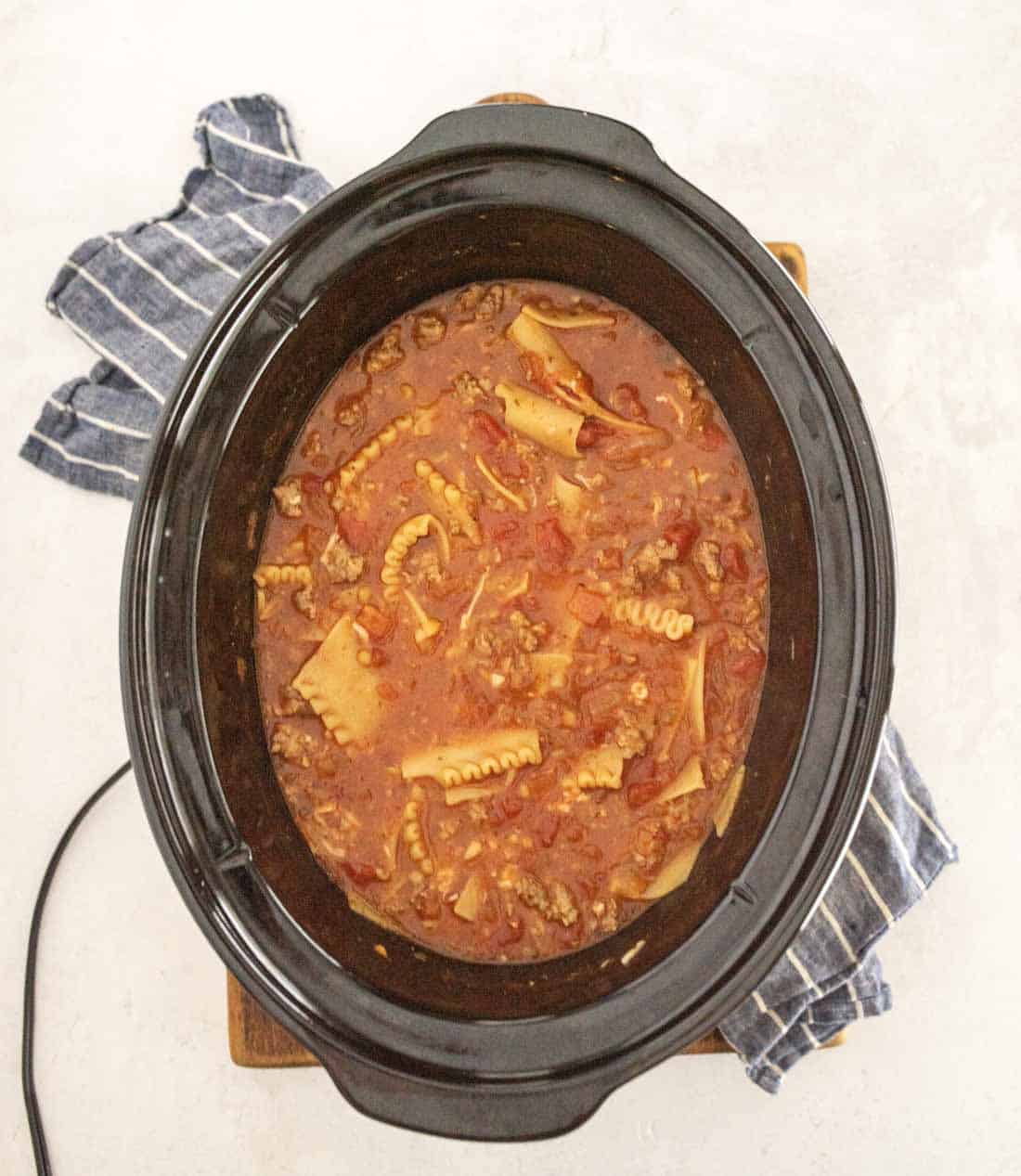A black slow cooker filled with lasagna soup in slow cooker, featuring broken lasagna noodles, ground meat, and rich tomato sauce, sits on a wooden board with a blue striped cloth nearby.