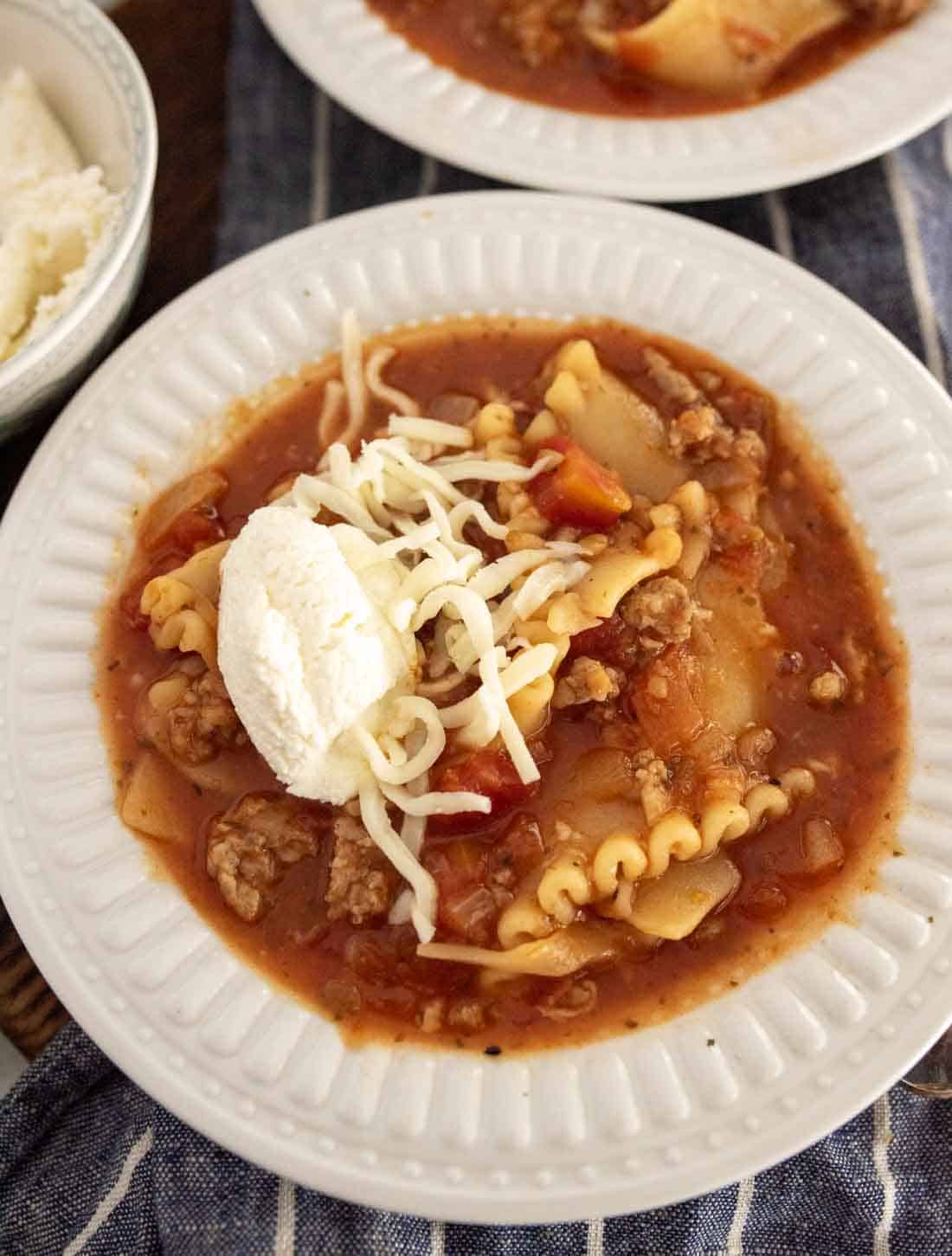 A bowl of lasagna soup in slow cooker, topped with shredded cheese and a dollop of ricotta, served in a white bowl on a striped cloth, with pasta, ground meat, and tomato sauce visible.