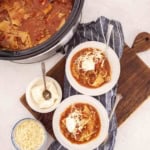A crockpot of lasagna soup in slow cooker sits next to two bowls of soup on a striped towel, each topped with cheese and sour cream. Nearby, bowls of shredded cheese and ricotta rest on a wooden board.