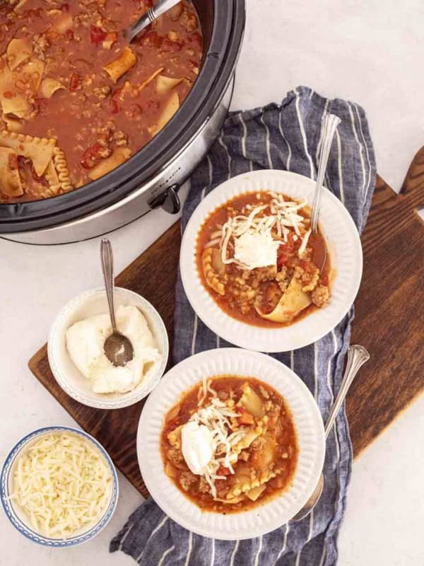 A crockpot of lasagna soup in slow cooker sits next to two bowls of soup on a striped towel, each topped with cheese and sour cream. Nearby, bowls of shredded cheese and ricotta rest on a wooden board.