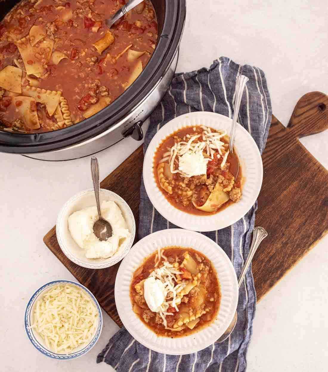 A slow cooker of lasagna soup in slow cooker sits next to two bowls of soup on a striped towel, each topped with cheese and sour cream. Nearby, bowls of shredded cheese and ricotta rest on a wooden board.