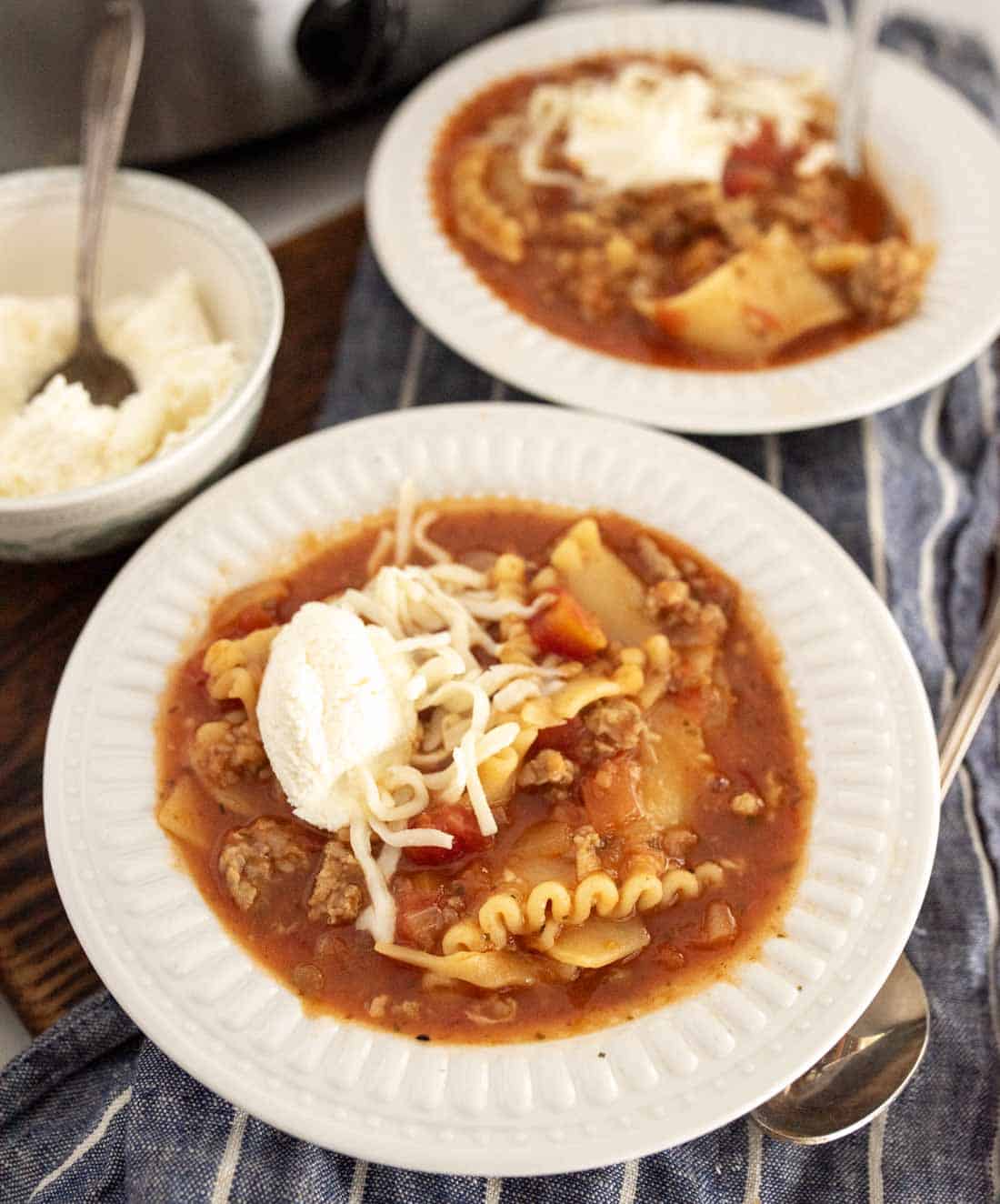 Two white plates filled with lasagna soup in slow cooker, topped with shredded cheese and a dollop of ricotta, sit on a blue striped cloth. A bowl of extra ricotta and a spoon are nearby.