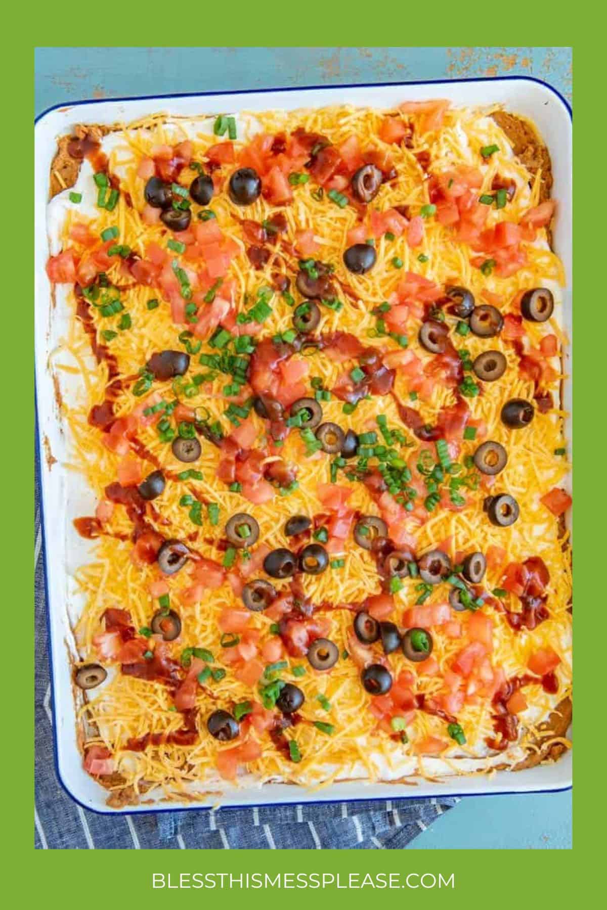 A casserole dish filled with a layered dip topped with shredded cheese, diced tomatoes, sliced black olives, green onions, and bacon bits&mdash;an irresistible choice for super bowl appetizers. Surrounded by a green border with BLESSTHISMESSPLEASE.COM at the bottom.