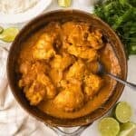 A skillet filled with chicken curry with chicken thighs coated in thick, orange sauce. Sliced limes, fresh cilantro, a bowl of white rice, and a beige towel surround the skillet on a white surface.