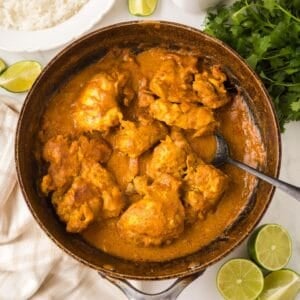 A skillet filled with chicken curry with chicken thighs coated in thick, orange sauce. Sliced limes, fresh cilantro, a bowl of white rice, and a beige towel surround the skillet on a white surface.